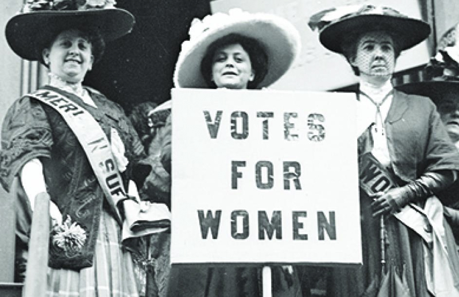 Three suffragists with sashes and a 'Votes for Women' placard